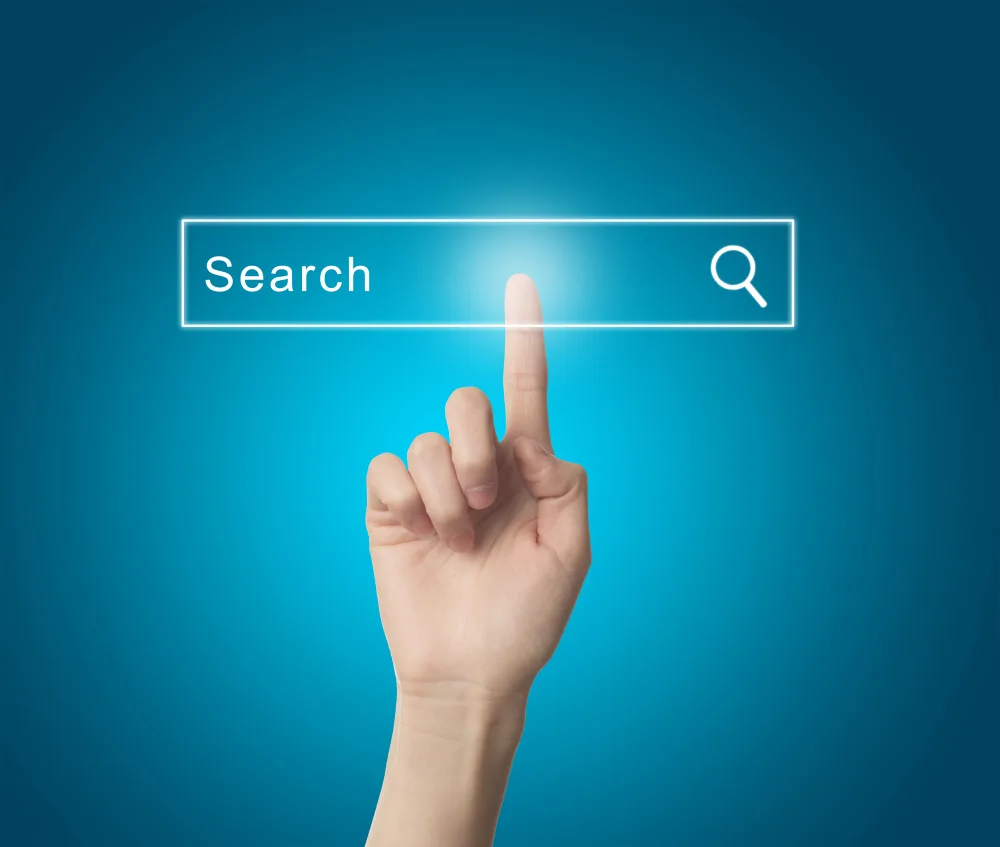صورة فوتوغرافية مقربة ليد بشرية تمتد من الأسفل لترتفع بسبابتها وتلمس شريط بحث رقمي مضيء ومستطيل يحتوي على كلمة "Search" باللغة الإنجليزية وأيقونة عدسة مكبرة على اليمين، حيث تظهر بؤرة ضوء بيضاء مشعة عند موضع اللمس، على خلفية زرقاء سادة متدرجة الإضاءة.